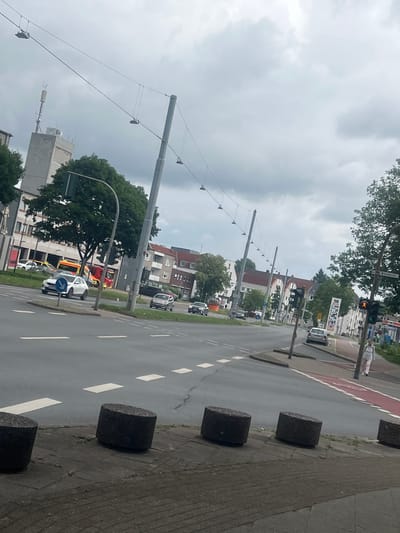 The image presents a tilted, daytime street view in Herne, Germany, under an overcast sky with heavy clouds. The scene features a multi-lane asphalt road with white dashed and solid lane markings, adjacent to a sidewalk partially covered by a red/brown path. Dark grey cylindrical bollards are spaced along the edge of the sidewalk.
Vehicles populate the road, including several cars, a white police car with blue markings, and a red and yellow emergency vehicle, possibly a fire truck or rescue vehicle, visible further down the street. A solitary pedestrian is seen walking on the sidewalk in the distance. Traffic lights are present, with one clearly displaying an amber/orange signal.
The street is lined with multi-story buildings exhibiting typical European architectural styles, including tiled roofs. Green trees with dense foliage are interspersed along the road and around the buildings. Overhead utility wires are strung between tall poles that line the street.
Visible text includes "Wiescherstr." on a street sign, providing a clear location cue. Other signs display "SB" and "Autoteile," likely indicating an auto parts store. Another sign with "SB" and numerical values (possibly fuel prices) suggests a petrol station or convenience store. A blue circular sign with a white arrow indicates a traffic directive.