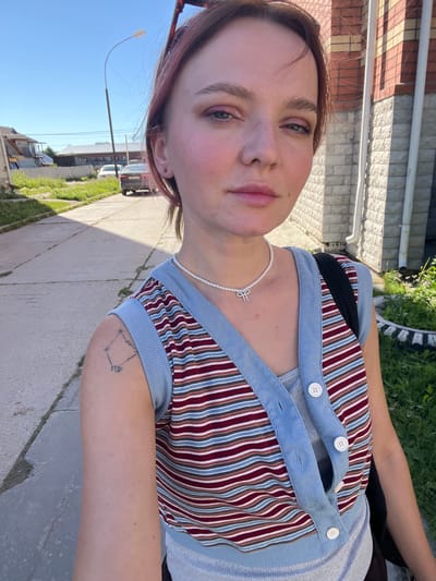 This is a portrait of a young person, taken outdoors in what appears to be a residential area. The subject is facing the camera, with their head tilted. They have light skin, with some pink blush on their cheeks. Their eyes are open, and they have light-colored eyebrows. Their lips are closed. Their hair is short and appears to be a light brown with some pink highlights.
The subject is wearing a striped top with blue and red stripes, layered over a grey top. They are also wearing a pearl necklace with a small, silver charm. A tattoo is visible on their upper arm, which appears to be a diamond shape with some symbols inside.
The background consists of a sidewalk, some grass, and a building with brick and stone accents. The lighting suggests it is daytime, likely sunny, given the shadows. The blue sky is visible in the upper portion of the image. The overall impression is casual and relaxed, possibly a candid shot. There is no visible text in the image.