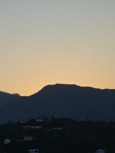 An image shows a mountainside at dusk, set against a gradient sky of pale yellow and light gray. A faint contrail streaks across the sky. The mountainside is dense with trees, giving way to visible structures on the lower slopes. The overall impression is of a tranquil, fading light over a natural landscape with hints of human presence.
