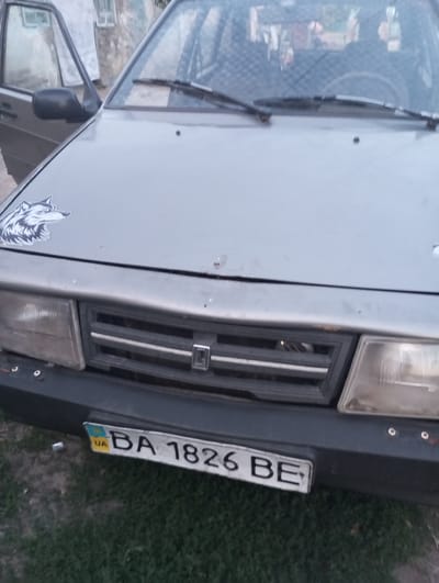 A gray sedan is parked on a grassy area in front of a building with white siding. The car's hood features a sticker of a wolf's head. The front grille has a horizontal design with a small emblem in the center. A license plate is visible on the lower front bumper, reading "UA BA 1826 BE". The car appears to be outdoors, with daylight illuminating the scene. The building in the background has windows. The overall condition of the car suggests it is an older model.