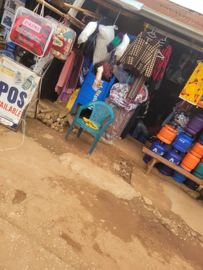 This is a photograph of an outdoor market stall in Tangu, Nigeria. The stall is filled with various goods, including stacks of fabric, bedding, and propane gas cylinders. On the left side of the frame, several blankets or comforters are neatly folded and displayed. One is red with a black and white camouflage pattern and "ESKIMO SUPER SOFT BLANKET" is printed on it. Another is a darker red with a patterned design and the word "KIMO" is visible. Above these, more folded textiles and some pillows are stacked. In the center of the stall, a turquoise plastic chair is placed, with a yellow cloth draped over its back. Behind the chair, a collection of brooms or brushes is tied together and resting on the ground.

To the right of the chair, a rack holds numerous propane gas cylinders in various colors: purple, orange, and blue. Some of these cylinders have branding visible, such as the letter "A" and what appears to be an Arabic script. The market stall appears to be set up along a dirt or dusty ground. The lighting suggests it could be daytime, likely morning or afternoon, with natural light illuminating the scene. The overall impression is one of a busy, functional marketplace. A poster or sign near the bottom right corner shows the words "POS AVAILABLE".