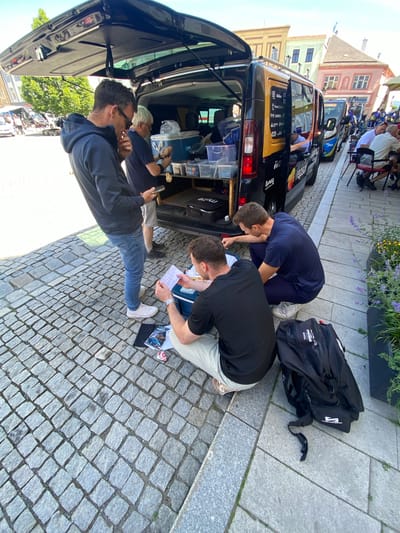 The image captures an outdoor scene in Uničov, Czechia, characterized by a group of men gathered around the open trunk of a dark-colored van. The van is parked on a cobblestone street, lined with parked cars in the background and historic buildings. Two men are bending over, examining papers, possibly maps or documents. One man in a blue top is leaning into the van. Another man is standing while using a cell phone. A black duffel bag sits on the cobblestone next to the van. The sky is sunny, indicating it's daytime. The presence of team bags and the activity around the van suggest that it is a support vehicle for an event, possibly a race.
