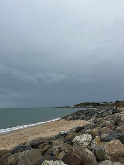 The image depicts a stony beach in Courtown, Ireland under a cloudy sky. The sea is a muted green, with gentle waves lapping onto the sandy shore. A prominent rock breakwater lines the edge of the beach, composed of large, irregular grey and brown stones. In the distance, along the coastline, a small cluster of houses can be seen nestled on a green hill. The sky is overcast with thick, dark clouds, suggesting impending rain or a storm. The overall scene is one of a rugged coastal landscape.