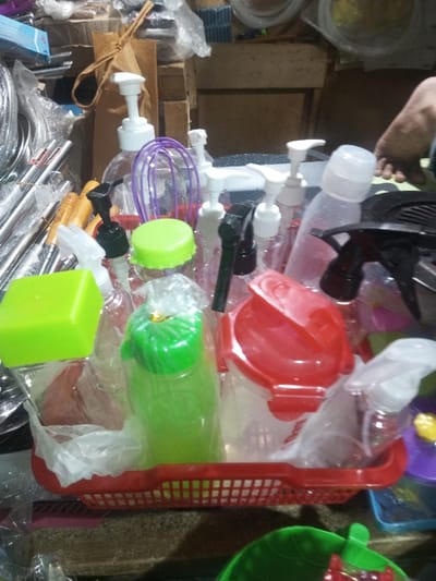A red plastic basket overflows with various plastic bottles, containers, and spray nozzles. Some have bright green, red, and clear lids. There are multiple pump-style dispensers and trigger sprayers. The containers are packed together, some wrapped in clear plastic. Metal kitchen utensils are visible to the left. A bare foot is partially seen to the right of the container.