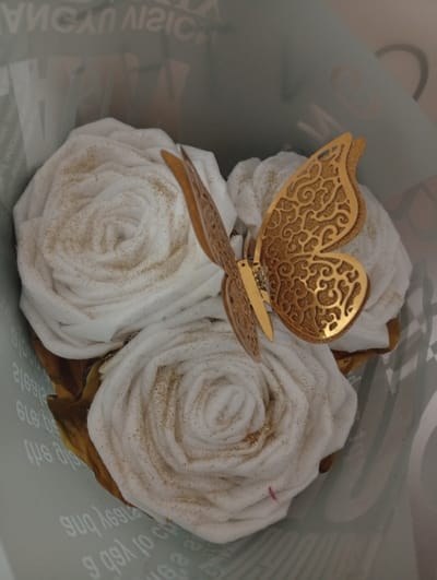 In Seýdi, Turkmenistan, a close-up, eye-level shot captures a decorative arrangement of three white, rose-shaped objects, possibly made of fabric or paper, dusted with gold glitter. A metallic gold butterfly ornament with intricate cut-out designs is positioned prominently on top of the arrangement. The objects are contained within a light blue paper bag, which has some text printed on its interior. The background is blurred, suggesting an indoor setting. The lighting is soft, casting gentle shadows. The scene is static, with no discernible action taking place.