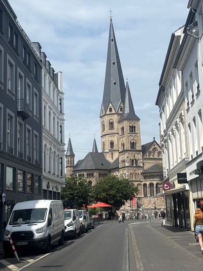 The image shows a street scene in Bonn, Germany, with a large Romanesque cathedral in the background. The cathedral features twin spires and intricate stonework. The street is lined with buildings on both sides, with shops on the right and residential or office buildings on the left. Several white vans are parked along the street on the left. People are visible walking on the sidewalk and crossing the street. The sky is partly cloudy, and the day appears to be bright and sunny. The overall scene depicts a lively urban environment with historical architecture. The prominent cathedral is likely the Bonn Minster.