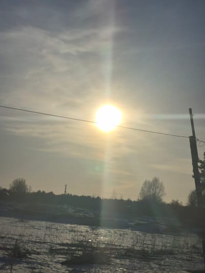 The image shows a winter scene in what appears to be the rural outskirts of a city. The sky is overcast, with the sun shining brightly through a thin layer of clouds, creating a strong lens flare. A utility pole with wires is visible in the upper left portion of the frame. The landscape is covered in snow. A line of trees is visible in the background. The location could be Дулесово, Russia.