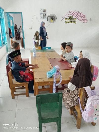 The image depicts a classroom scene within TK Uswatun Hasanah, a kindergarten located in Jagapura Kulon, Indonesia, specifically captured at 07:32 on July 14, 2025. The environment features white walls, a light-colored tiled floor, and blue-framed windows on the left. A wall-mounted fan and a "TK USWATUN HASANAH" wall decoration are visible on the back wall.
Approximately seven children are present, mostly seated at small, light-colored wooden tables and chairs. Two children wear Islamic peci (skullcaps); one boy in a black peci has "M. Arvan" written on it and a red bag on his lap. Several girls wear hijabs or headscarves. On the tables, two plastic water bottles with carrying straps are visible, one blue and one purple. A light purple backpack with a cartoon character hangs from a chair in the foreground.
In the background, a woman in a blue dress and patterned hijab stands near the children's tables, holding a dark object. Through an open blue-framed doorway on the left, two additional adults—a woman in a patterned dress and a partially visible man—are present in an adjacent room. The children appear to be settled, with one child looking directly at the camera.