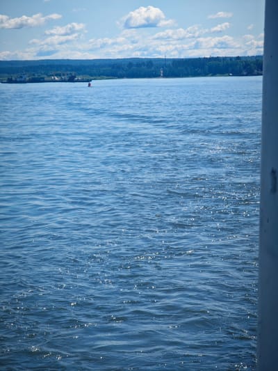 The image shows a body of water with a distant shoreline lined with trees and some buildings. The sky is blue with scattered white clouds. The water is rippled and reflects the sunlight, creating sparkling highlights. A red buoy is visible in the water, as well as a barge or boat further in the distance near the shore. The right side of the image is partially obstructed by a vertical, light-colored object, possibly a railing or wall of a vessel. The scene is serene and natural, evoking a sense of a peaceful day on the water. The provided context suggests the location is Krutoy Log, Russia.