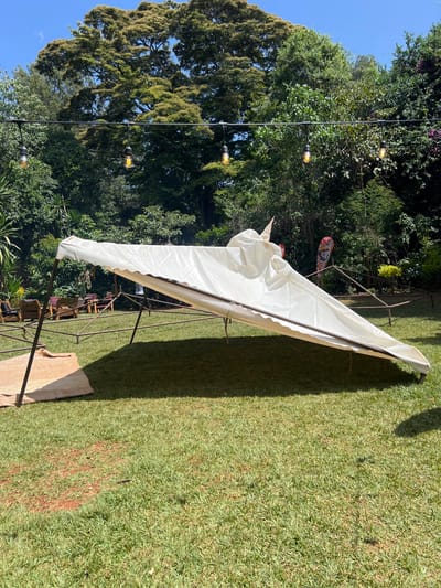 This media file captures an outdoor scene in what appears to be a garden or event space in Mururiini, Kenya. The dominant subject is a white canopy or tent structure, elongated and triangular in its visible section, supported by dark metal poles. The canopy provides shade, casting a distinct shadow on the vibrant green grass below.
The setting is lush and green, featuring extensive, well-maintained grass and numerous large, mature trees that form a dense canopy in the background and along the left side of the frame. A string of decorative Edison-bulb lights is strung horizontally across the upper-left quadrant, indicating that the space is also prepared for evening use. In the distance, beneath the tent, faint glimpses of what appear to be chairs and tables suggest a gathering or dining area. A piece of unfinished wooden board or slab rests on the grass near the front-right leg of the canopy structure. A small, colorful decorative flag or banner is visible among the trees in the mid-background, to the right of the string lights.
The time of day is bright, with clear skies and strong sunlight, as evidenced by the distinct shadows cast by the canopy and trees. The overall impression is one of a peaceful and inviting outdoor venue, likely prepared for an event or casual relaxation. There is no visible text in the image.