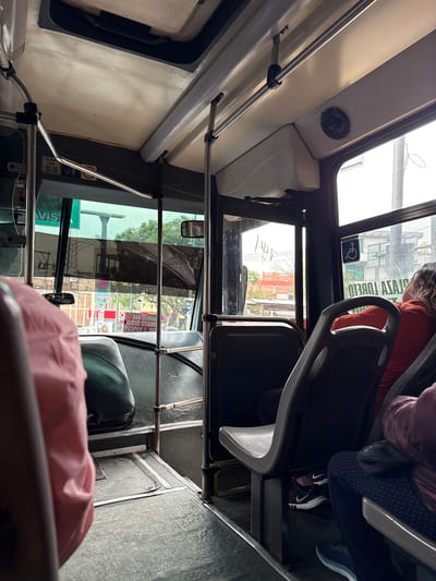 The image shows the interior of a bus with passengers. The perspective is from a seat looking towards the front and side of the bus. There are metal poles for support and several seats visible. Outside the window, trees, buildings, and signs can be seen, suggesting an urban environment. One sign visible through the window reads "TIZAPAN II IND." and another indicates "PLAZA LORETO". A person in a reddish-orange shirt is seated, facing away from the camera. Another passenger's pink clothing is in the foreground. The floor of the bus is visible and appears to be made of a textured metal. The lighting suggests it is daytime.