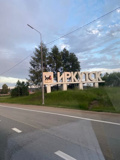 A welcome sign to Irkutsk, Russia, stands on the side of a road. The sign consists of large, white, three-dimensional letters spelling out "ИРКУТСК" (Irkutsk). To the left of the letters is a crest with a black sable holding a red bow. The sign is situated on a grassy hill, with trees behind it. A lamppost stands to the left of the sign. The road in front of the sign has white lane markings. The sky is partly cloudy. The overall scene suggests the outskirts of a city or town.