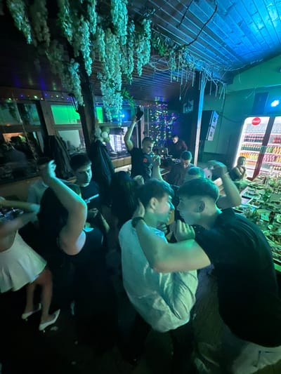 The image shows a gathering of people, predominantly young adults, indoors. The location appears to be a bar or restaurant, based on the presence of a beverage refrigerator and decorative elements like faux cherry blossom branches. The lighting is a mix of neon and ambient, with strong blue and green hues. Several individuals are interacting closely, while others are seated or standing nearby. The overall atmosphere seems lively and social. Specific details regarding the city of Panagyurski kolonii, Bulgaria, are not evident within the image itself.