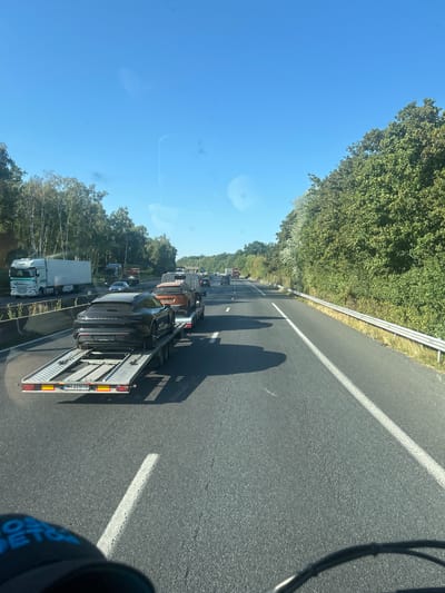 The image shows a highway in France with traffic. In the foreground, a car carrier trailer is visible, carrying a black Porsche. Behind it, an orange SUV is also on the trailer. The highway is bordered by trees on the right and left. The sky is clear and blue. The location is identified as Thiers-sur-Thève, France.
