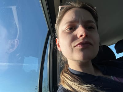 A close-up selfie of a young woman with light skin and brown eyes, likely in her late teens or early twenties. Her hair is pulled back into a low ponytail. She is wearing a dark blue jacket with a hint of pink underneath. The woman is inside a car; a car window is visible to the left, and a reflection of her ear can be seen in the window glass. A clear blue sky is visible outside the window. The image is taken from a low angle, making her face appear dominant. The scene does not include any information to determine the location in Skagen, Denmark.