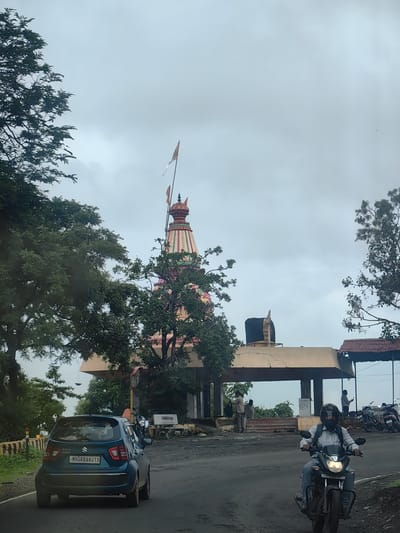 This is a full-shot, eye-level, outdoor photograph taken on a cloudy day on a paved road in Mohegaon, India. In the foreground, a blue Suzuki Ignis car is visible from the rear, driving away from the camera. The car has a license plate that reads "MH46BA813". To the right of the car, a person wearing a helmet is riding a black motorcycle, also driving away from the camera. The motorcycle has a license plate that reads "SMR 26BB 1620".
In the midground, there is a temple with a colorful shikara, or spire, topped with flags. Several people are standing near the temple entrance, some appearing to be looking at their phones. A covered walkway or structure is present in front of the temple.
The background consists of lush green trees and the overcast sky. The overall scene depicts a roadside view of a temple in a rural or semi-urban setting.