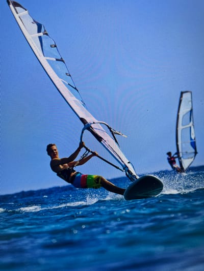 The image shows a windsurfer performing a trick in the water. The windsurfer is upside down, holding onto the boom of the sail with both hands. The sail is white with blue accents. The board is dark green. The water is blue and choppy. In the background, another windsurfer is visible in the distance. The location is possibly in the city of Bacong, Philippines.