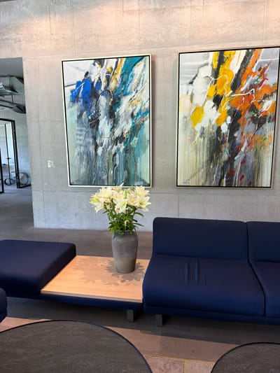The image shows an indoor scene with two abstract paintings hanging on a gray wall, a navy blue sofa, a small wooden table with a gray vase containing white lilies, and two round black tables. Below the paintings are visible parts of a metal structure, perhaps part of a rack or stand. The location is Chkalov, Armenia, given the prompt.