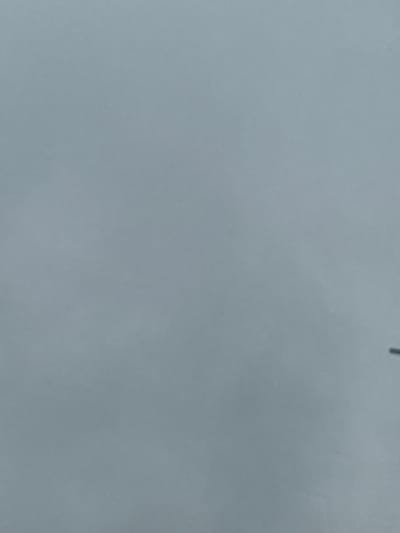 The image displays a close-up view of a uniformly colored surface, predominantly a light, muted blue-gray or slate gray hue. The surface appears matte with a subtle, fine-grained texture, suggesting it could be a painted wall, a large piece of paper, or fabric. In the original image, and visible in some cropped hints, a very faint, thin, dark vertical line segment is present near the top center-right edge of the frame. This line is indistinct and could be a stray mark, a thin wire, or a string. There are no discernible people, specific objects (other than the surface itself and the faint line), or visible text. No activities or events are taking place. The image does not provide any visual cues regarding the time of day, weather, or a specific location within Bashkibash, Russia, which was provided as context.