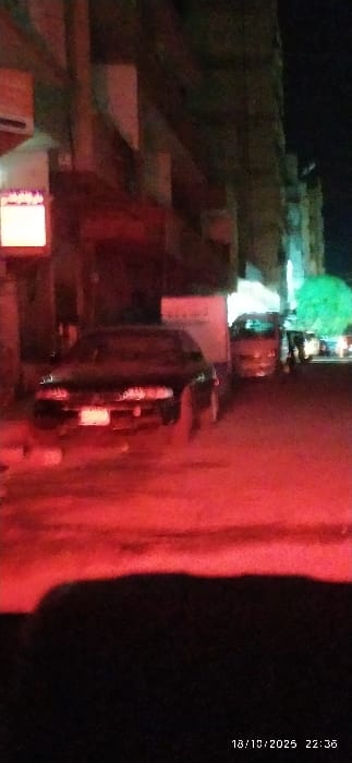 The image depicts a nighttime street scene in the city of Al-Kiswa, Syria. A dark-colored sedan-type vehicle is positioned in the foreground, facing away from the viewer, its rear illuminated by a prominent red light source originating from the immediate environment. The red light casts a significant glow across the foreground and midground. To the right, further down the street, a lighter-colored vehicle, possibly a van or small truck, is partially visible. Buildings with multiple stories line both sides of the street, exhibiting some signs of lighting or commercial activity. A bright, rectangular white sign is visible on the left side of the street, attached to a building. Further in the distance on the right, a distinctive green light source is visible, suggesting illuminated foliage or a commercial sign. The street surface is indistinct due to lighting and image quality. No individuals are discernible within the frame. The timestamp 18/10/2025 22:36 is present in the lower right corner.