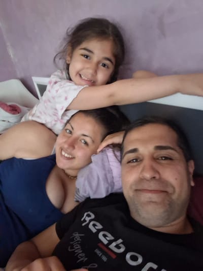 In a bedroom in Novachene, Bulgaria, a family is posing for a selfie. A young girl is positioned at the top, her head resting on her mother's shoulder. The mother is in the middle, smiling broadly, with her arm around the girl. The father is at the bottom, also smiling, taking the photo. All three individuals appear to be happy and relaxed. The background is a soft purple wall. The overall impression is one of warmth and family connection.