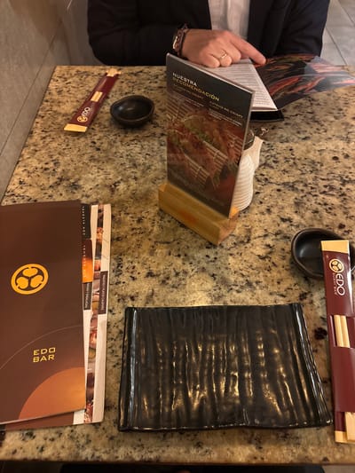A person's hand, wearing a gold ring, is shown holding open a menu at a table. The table appears to be made of granite. In front of the menu, there is a small black bowl and a set of chopsticks in a maroon wrapper with the "EDO SUSHI BAR" logo. To the right of the bowl is a rectangular black plate. Further to the right, another menu is visible, which is dark brown with the "EDO BAR" logo. In the background, there are more menus stacked. The setting appears to be a restaurant, possibly an Edo Sushi Bar in Magdalena del Mar, Peru. The lighting suggests it could be daytime. The overall scene conveys a casual dining experience. Visible text includes "EDO SUSHI BAR", "EDO BAR", "NUESTRA RECOMENDACIÓN", "PAKAGES Y PLATOS CALIENTES", "SIN ALCOHOL", and "CON ALCOHOL".