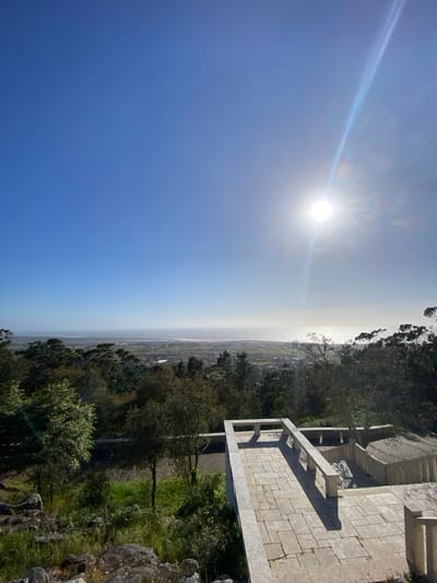 The image shows a landscape view from a high vantage point, likely in Esposende, Portugal. The sun is prominent in the clear blue sky, with a visible lens flare. Below the sky, the land stretches out towards the coast, with the ocean or a large body of water visible in the distance. The view includes a mixture of urban and rural areas, with buildings and fields. In the foreground, there is a stone structure, likely a viewing platform or part of a building, with a stone floor and a railing. Lush greenery, including trees and shrubs, surrounds the structure and extends down the slope. The picture seems to be taken in the daytime.