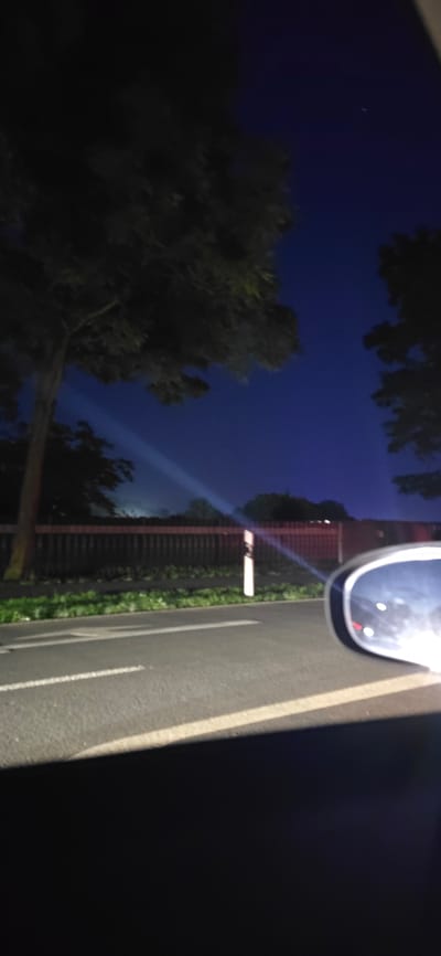 The image depicts a nighttime scene viewed from a vehicle, likely a car, driving along a road. The sky is a deep, dark blue, characteristic of twilight or a clear night. In the foreground, the right side of the image shows a car's side mirror, reflecting lights from the vehicle's interior or the surrounding environment. The road surface is visible, with white lane markings indicating it's a paved road. To the left and extending into the background, trees with dark foliage frame the scene. A light beam or ray cuts across the sky from the left towards the center, suggesting a light source from beyond the frame or a spotlight. Behind the trees and a wooden fence, a subtle glow of light suggests distant city lights or buildings. The overall impression is that of a drive through a semi-urban or rural area at night, possibly near Neustadt-Glewe, Germany, given the context provided.