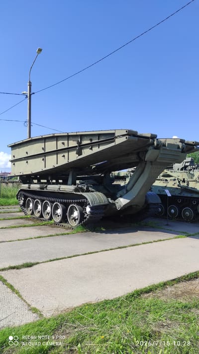 This image depicts an outdoor display of military vehicles, likely at a museum or memorial site, in Лашанскі сельскі Савет, Belarus. The primary subject is a large, olive-drab military engineering vehicle, which appears to be a bridge-laying tank. It is positioned on a paved surface surrounded by grass. The vehicle has a prominent, folded bridge structure mounted on top of its tracked chassis. The chassis itself features multiple road wheels and a robust track system. Behind this vehicle, another olive-drab military vehicle, possibly a tank, is partially visible. The background shows a clear blue sky with some power lines and a lamppost. The scene is captured on a bright, sunny day. The lower right corner of the image contains a timestamp indicating "2026/7/15 10:28" and text suggesting it was captured by a "REDMI NOTE 9 PRO AI QUAD CAMERA."
