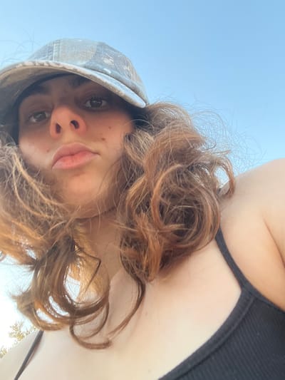 The scene is located in the Brener Regional Council, Israel. The image depicts a close-up, low-angle portrait of an individual, appearing to be a young female, occupying the majority of the frame. She has light skin, dark eyes, and medium-length wavy reddish-brown hair. She is wearing a camouflage-patterned baseball cap, visible on her head, and a black ribbed tank top with thin shoulder straps. Her face is directed towards the camera with a neutral expression, lips slightly pursed. The background consists primarily of a clear, bright blue sky. A small amount of green foliage, indicative of trees or bushes, is visible in the lower left corner of the frame. The lighting suggests natural daylight.