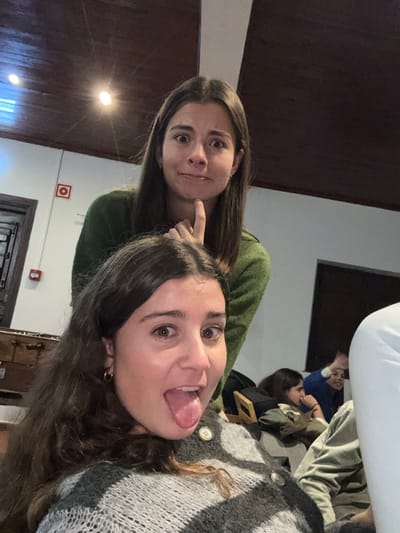 Two young women pose for a photo. One, in an olive green shirt, stands behind the other, touching her chin with a playful expression. The second woman sticks her tongue out, wearing a gray sweater with large buttons. In the blurry background, additional people sit at a table. The interior space features dark wood paneling and a white ceiling. A red fire alarm and doorway are visible in the background.