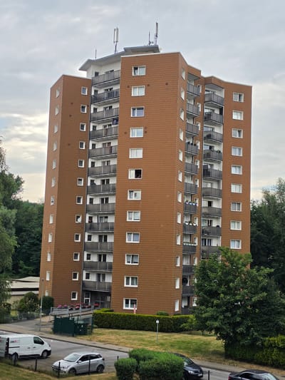 This image shows a tall, brown apartment building located in Oberhausen, Germany. The building has many balconies with dark railings, and numerous windows of various sizes. The exterior of the building appears to be covered in a textured brown material, possibly shingles or siding. The roof of the building has several antennas and a flat section.

In front of the building, there is a road with a white van and a silver car parked. There is also a black car further down the road. Green hedges and grass are visible in the foreground and surrounding areas. Trees are also present, with some appearing to be very close to the building on the left side, and a large tree on the right side of the image. The sky is overcast.

There are at least two people visible on balconies of the apartment building, one appearing to look out of a window on a lower floor and another on a higher floor. Another person is visible on a balcony in the middle section of the building, looking towards the camera. Several balconies have planters with flowers.