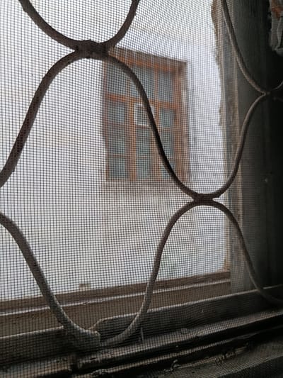 The image shows a close-up view through a window screen, revealing a blurred outdoor scene. The foreground is dominated by the mesh of the screen and decorative, curved metal bars, which are somewhat rusty. Beyond the screen, a window with wooden frames is visible, also out of focus. The overall impression is one of looking out from an interior space into a subdued, possibly overcast environment. There are no people or specific activities depicted. The location context indicates this is from Navruz, Uzbekistan, suggesting a possible cultural or seasonal relevance, though this is not visually apparent. The time of day and weather are not clearly discernible due to the blurriness and lack of distinct light sources. No text is visible in the image.