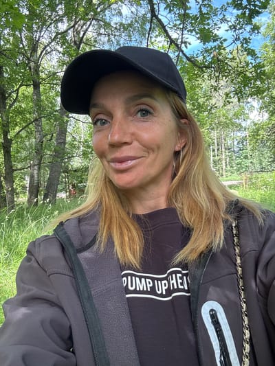 A woman is taking a selfie in a wooded area with lush green grass and trees. She is wearing a black baseball cap, a dark jacket over a black sweatshirt with white text that reads "PUMP UP HEI", and large hoop earrings. The woman has blonde hair and is smiling at the camera. The background shows a sunlit forest with dappled light filtering through the leaves.