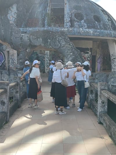 The image captures a group of individuals, predominantly adult women, on a paved pathway or bridge in Kim Long Ward, Vietnam, in front of a large, decaying concrete structure. This structure is identified as part of the abandoned Ho Thuy Tien water park, specifically featuring a prominent, weathered dome and an integrated dragon-themed architectural element.
The central subject is the dilapidated concrete edifice, which includes a colossal, scaled dragon's head and body wrapping around a lower section and supporting a large, circular dome above. The entire structure is heavily covered in multi-colored graffiti tags and abstract spray-painted designs. Circular openings are visible on the dome, which shows signs of significant weathering and decay, including some exposed rebar and sparse vegetation growing from crevices.
On the light-colored tiled pathway in the foreground, approximately ten to twelve individuals are present. Most are dressed in casual attire, including white tops, a mix of black skirts, maroon skirts, and blue jeans, along with various hats such as white baseball caps and light-colored bucket hats.
* One woman on the left, wearing a white baseball cap, white short-sleeved top, black midi skirt, and light-colored sneakers, stands with a red and white striped tote bag, looking back towards the viewer.
* In the middle, a woman wearing a bucket hat, white long-sleeved top, black midi skirt, and light-colored sneakers with brown socks, extends her right arm forward, pointing towards the structure. She carries a light brown crossbody bag.
* To her right, another woman, wearing a white baseball cap, sunglasses, a white "Life Says" t-shirt, blue jeans, and white sneakers, holds a smartphone up, appearing to photograph or video the scene. She carries a white crossbody bag.
* Other individuals are clustered around, observing the structure or engaging in conversation. One person is partially visible in an archway of the structure, looking towards the group.
The scene is under bright daylight, evidenced by distinct shadows cast on the pathway, suggesting the sun is positioned to the upper right. Concrete railings with decorative, undulating patterns line both sides of the pathway. The overall environment conveys a sense of abandoned, yet still visited, architectural ruins.