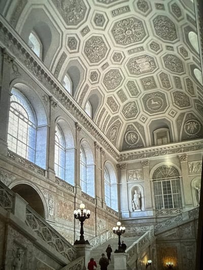 This image depicts a grand interior architectural space, located in the city of Senokos, Bulgaria. The scene is dominated by a majestic, bifurcated marble staircase with ornate balustrades featuring intricate geometric carvings. The walls are adorned with numerous tall, arched windows, each with multiple panes, extending through multiple levels and admitting significant natural light. The high ceiling is elaborately coffered, primarily with hexagonal and some rectangular panels, each containing detailed bas-reliefs depicting mythological figures, animals such as a deer, human forms, and botanical motifs. The architectural style includes pilasters, decorative friezes, and cornices.
On the upper levels, several niches are visible within the wall structure; one prominently displays a white classical statue of a standing male figure. The staircase is illuminated by two large, black, multi-armed lampposts, each with multiple globe lights, positioned on the balustrades at a lower landing.
Several individuals are present on the staircase. In the foreground on the left, one person wears a red top, and another wears a dark jacket, both ascending. Further up the left branch of the staircase, an individual in a dark coat stands. On the right branch, another person in a dark coat is visible near a lamppost, also appearing to ascend.