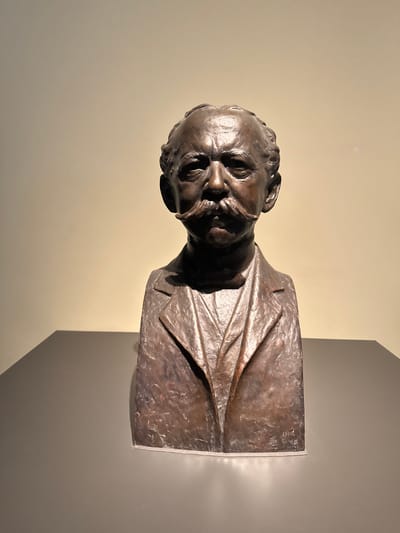 A bronze bust of a man with a prominent mustache stands on a dark grey pedestal against a light beige wall. The bust depicts the man from the shoulders up, wearing a suit jacket and a collared shirt with a tie. His hair is styled back, and he has a stern expression. There appears to be a signature and date inscribed on the right side of the bust. The lighting creates shadows on the wall behind the sculpture, suggesting it is indoors, likely in a museum or gallery setting in Bern, Switzerland.