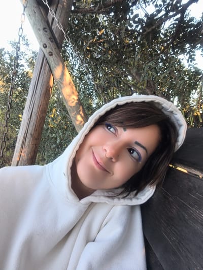 An outdoor, eye-level photograph captures a young adult female against a backdrop of wooden structures and foliage in Shaalvim, Israel. The female, positioned in the lower-left portion of the frame, has short dark brown hair visible under an off-white hooded sweatshirt. Her head is tilted slightly upward and to the left, displaying a gentle smile. She has dark eyes with blue eyeliner on the lower lash line, defined eyebrows, a small stud piercing above her left eyebrow, and a gold hoop nose ring in her left nostril. A small dark mark is present above her upper lip. She leans against a dark, weathered wooden surface that occupies the right side of the image. The background features two large wooden posts, likely part of an outdoor structure, with a metal chain hanging vertically on the left. One diagonal wooden post shows circular indentations. Dappled sunlight creates bright spots on the wooden elements. Dense green foliage and tree branches with visible green pods fill the upper background, with patches of bright sky visible through the canopy.