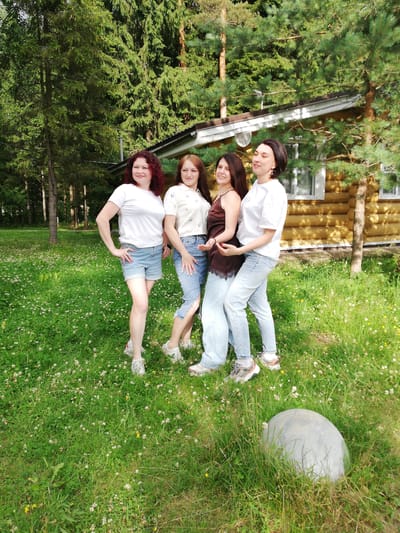 Four women are standing in a grassy area with a log cabin in the background. The woman on the far left has curly red hair and is wearing a white t-shirt and denim shorts. The second woman from the left has straight brown hair and is wearing a light-colored shirt and jeans. The third woman from the left has dark brown hair and is wearing a dark brown camisole and light-colored pants. The woman on the far right has dark brown hair and is wearing a white t-shirt and light-colored pants. The log cabin has a white roof and windows. There are trees in the background. The ground is covered in green grass and white flowers. A large, round, gray object is in the foreground on the right. The image appears to have been taken in the city of Shapki, Russia.