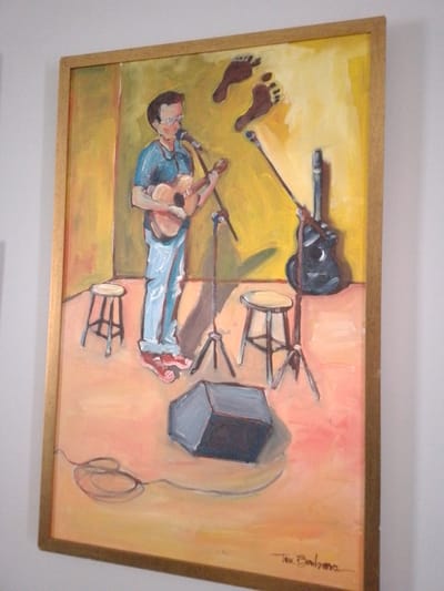 The image displays a vertically oriented, framed oil painting mounted on a plain white wall in Campo Grande, Brazil. The painting, rendered with visible brushstrokes, depicts a male musician performing on what appears to be a stage.

The central figure, a light-skinned male with short dark hair and glasses, stands slightly left of center. He is wearing a blue polo shirt, light blue jeans, and red shoes. He is actively playing a light-brown acoustic guitar and singing into a silver-colored microphone mounted on a black stand directly in front of him.

On the stage floor, two light-colored wooden stools are present: one to the musician's left and another to his right. A second black microphone stand is positioned on the right side of the stage, against which a black acoustic guitar leans vertically. In the foreground, a dark gray rectangular speaker or amplifier box rests on the stage floor, connected to a coiled cable visible in the lower-left portion of the painting.

The background of the stage consists of a yellow wall, with the floor painted in shades of orange and pink. Two stylized, dark brown human footprints are depicted on the upper-right section of the yellow wall, appearing to float above the scene.

The artist's signature, "Teca Balbina," is visible in dark script in the bottom right corner of the painting. The painting is enclosed within a simple, light-colored wooden frame.