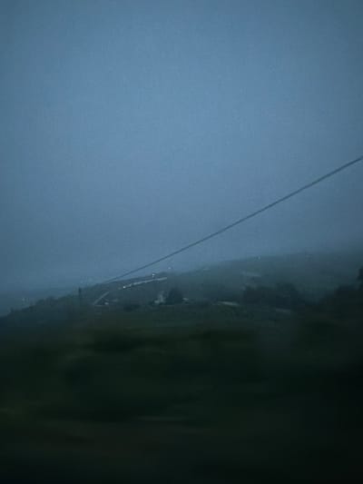 The image depicts an outdoor scene in Santo Stefano Belbo, Italy, likely taken during twilight or under heavy cloud cover. The foreground is dominated by an expanse of cloudy, grey-blue sky. A thin power line or wire stretches diagonally across the frame. The ground, possibly a hillside, is visible along the right side of the image, mostly obscured by dark vegetation and shadows. There are scattered lights indicating a built-up area or houses. The overall atmosphere suggests a muted and possibly misty environment.