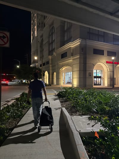 The image captures a nighttime urban scene, likely in Coral Gables, United States, showcasing a street and a commercial building. A person, viewed from behind, is walking on a sidewalk, pulling a black rolling shopping cart with a metal frame. They are dressed in a blue t-shirt and light-colored pants.
The prominent building on the left features a light-colored facade with classical architectural elements, including arches and cornices. Exterior lights illuminate its structure. Visible text on the building identifies "TRADER JOE'S" above one of the arched entrances, suggesting a grocery store. Below this sign, numbers "211" and "215" are visible, likely suite or address numbers, with "200 C" seen further down.
The sidewalk on the right is bordered by a raised planter filled with dense green bushes and smaller plants. Shadows from the foliage and the person are cast on the pavement due to artificial lighting. In the background, beyond the person and the building, a street is visible with distant streetlights and the red taillights of vehicles, confirming the time of day as night. A circular traffic sign with a red border and a crossed-out upward arrow, indicating "No Straight Through" or restricted access, is visible in the lower left corner. The weather appears clear.