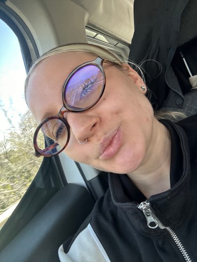 A close-up, high-angle selfie captured inside a vehicle depicts a female individual in Ula, Turkey. The subject has blonde hair, light blue eyes, and fair skin with visible blemishes. She is wearing tortoiseshell-patterned prescription glasses; the left lens exhibits a significant fracture pattern originating near the center and radiating outwards. A silver stud earring is present in her left earlobe, and a silver hoop nose ring pierces her septum. Her lips are pursed, forming a "duck face" expression. She is dressed in a black zippered jacket featuring a prominent silver zipper pull and a white stripe on the right sleeve. The vehicle's interior shows a white headliner in the upper left and a dark-colored panel below the window. Through the driver's side window, a clear blue sky is visible at the top, and blurred green foliage suggests an outdoor, possibly moving, environment. A dark, possibly fabric-covered object, is visible in the upper right background within the vehicle.