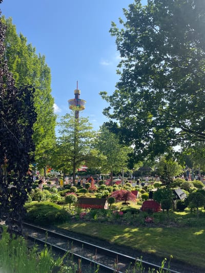 The image depicts a section of LEGOLAND Billund, Denmark, on a clear, sunny day. In the background, a tall tower with a colorful observation deck, branded "LEGO," stands prominently against the blue sky. The foreground features miniature LEGO models of buildings and landscapes, including red houses, a barn, and meticulously manicured trees and bushes. Several people are visible walking through the park, observing the displays. Railway tracks run along the bottom of the image, adding to the miniature theme. Lush green trees frame the scene, with dappled sunlight filtering through their leaves, creating a warm and inviting atmosphere.