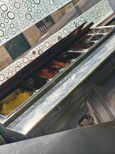 The image captures a food preparation station, likely inside a fast-food establishment in Moaña, Spain. The station features several stainless steel trays filled with various ingredients. One tray contains shredded cheese. Other trays appear to hold meat toppings, such as ham or bacon. The station is positioned by a window adorned with a circular pattern. Outside the window, a streetscape is visible, with buildings and a metallic pole. The environment suggests an urban setting.