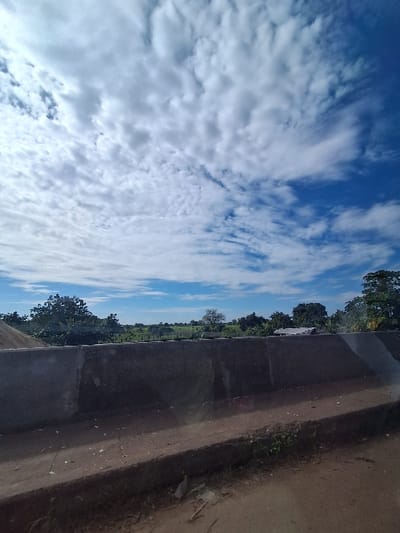 The image depicts a daytime, rural or semi-rural landscape in Kungana, Nigeria. The upper two-thirds of the frame features a blue sky with extensive scattered white cumulus and cirrus-like clouds. A dark gray concrete barrier, appearing weathered, traverses horizontally across the mid-section of the image, showing some surface reflections. The immediate foreground consists of an unpaved dirt surface with sparse green vegetation and scattered debris. Beyond the concrete barrier, a verdant landscape extends into the distance, characterized by numerous green trees, open fields, and faintly visible, partially obscured structures in the background, possibly buildings with light-colored roofs. No individuals or dynamic actions are present in the scene.