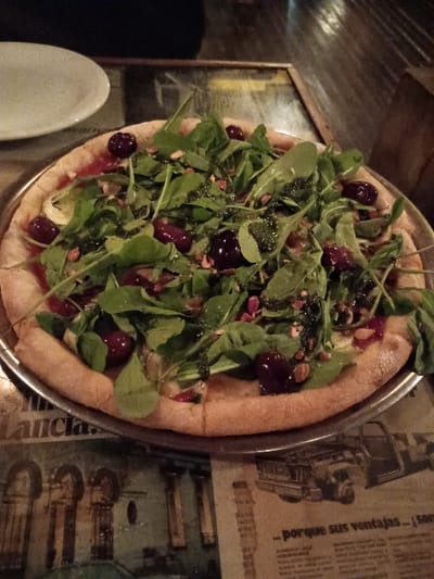 A pizza with a thin crust topped with arugula, olives, nuts, and a white sauce is sitting on a metal plate. The pizza is on a table with a newspaper underneath. The table is located in a restaurant, and the environment is dimly lit.