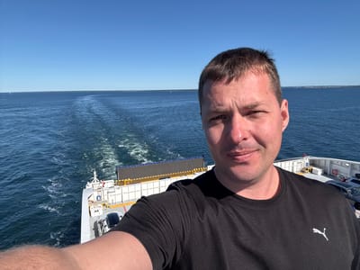 A man with short brown hair and a black t-shirt takes a selfie on the deck of a ferry. The ferry is moving through the sea, leaving a wake behind it. In the background, the water is blue and calm, with a clear sky above. The man is smiling at the camera. The ferry is white with some red and yellow accents. There are cars parked on the deck of the ferry. The scene appears to be taking place during the daytime. The location is the sea near Karlskrona, Sweden.