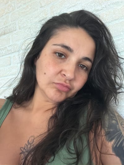A close-up, selfie-style shot captures a woman in her late 20s or early 30s with long, dark, wavy hair and brown eyes. She is wearing a green tank top, and a tattoo of a celestial design is visible on her left collarbone. Another large, dark tattoo covers her right arm. The woman's expression is a pout, with her lips pursed. She has light skin with some freckles and a mole on her left cheek. The background is a lightly textured, off-white wall. The image appears to be taken indoors, with soft lighting. There is no indication of the location being Ароматненское сельское поселение, Ukraine.