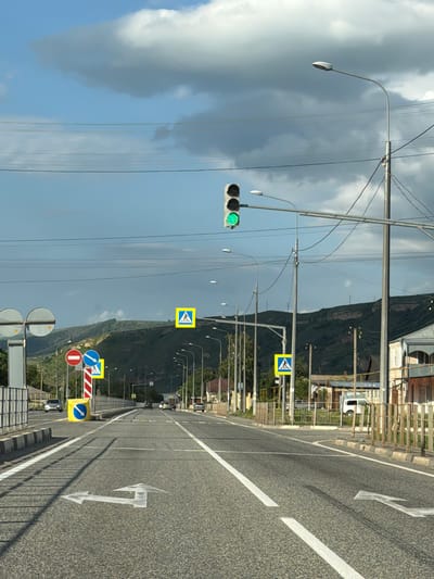 The image depicts a straight, paved road in Важное, Russia, with a green traffic light illuminated. On the left side of the road, there is a sidewalk bordered by a metal fence and some vegetation. Several traffic signs are visible, including a "no entry" sign, a blue circular sign with a white arrow indicating a turn, and a yellow square sign with a pedestrian crossing symbol. Further down the road, more pedestrian crossing signs and streetlights are present. The right side of the road features buildings and more metal fencing. The sky is partly cloudy with a mix of blue sky and grey clouds. The overall scene suggests a typical urban or suburban street environment.
